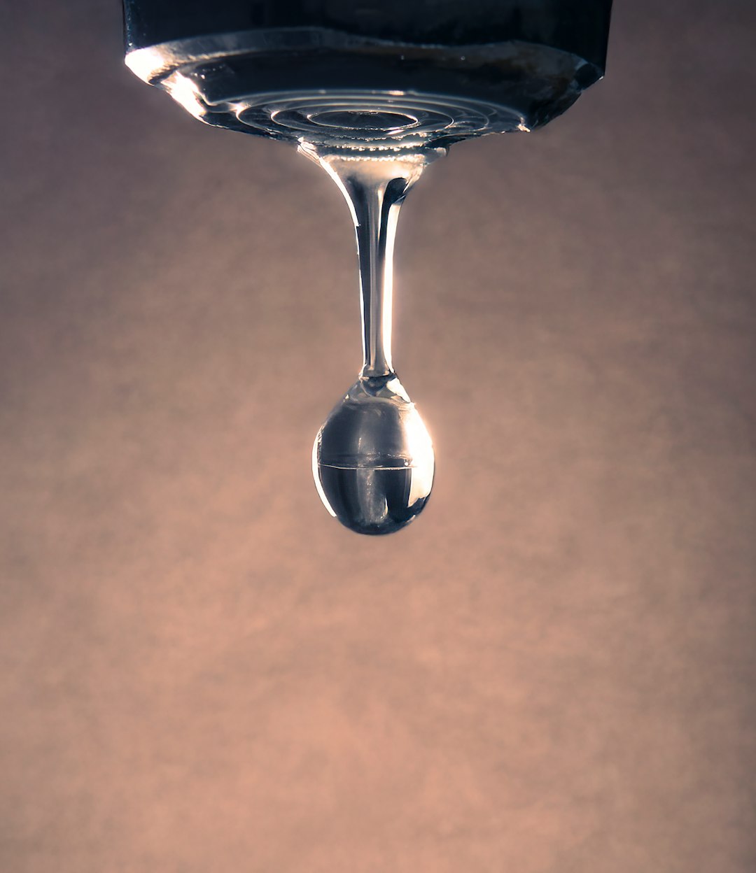 A high-resolution macro photograph capturing a single water droplet suspended in time, hanging from the bottom of a faucet. The image is artistically flipped upside down, emphasizing the tension and fragility of the moment just before the drop falls. Warm, diffused lighting in the background adds contrast and elegance, making it ideal for use in creative projects focused on themes like time, fragility, nature, or calmness.